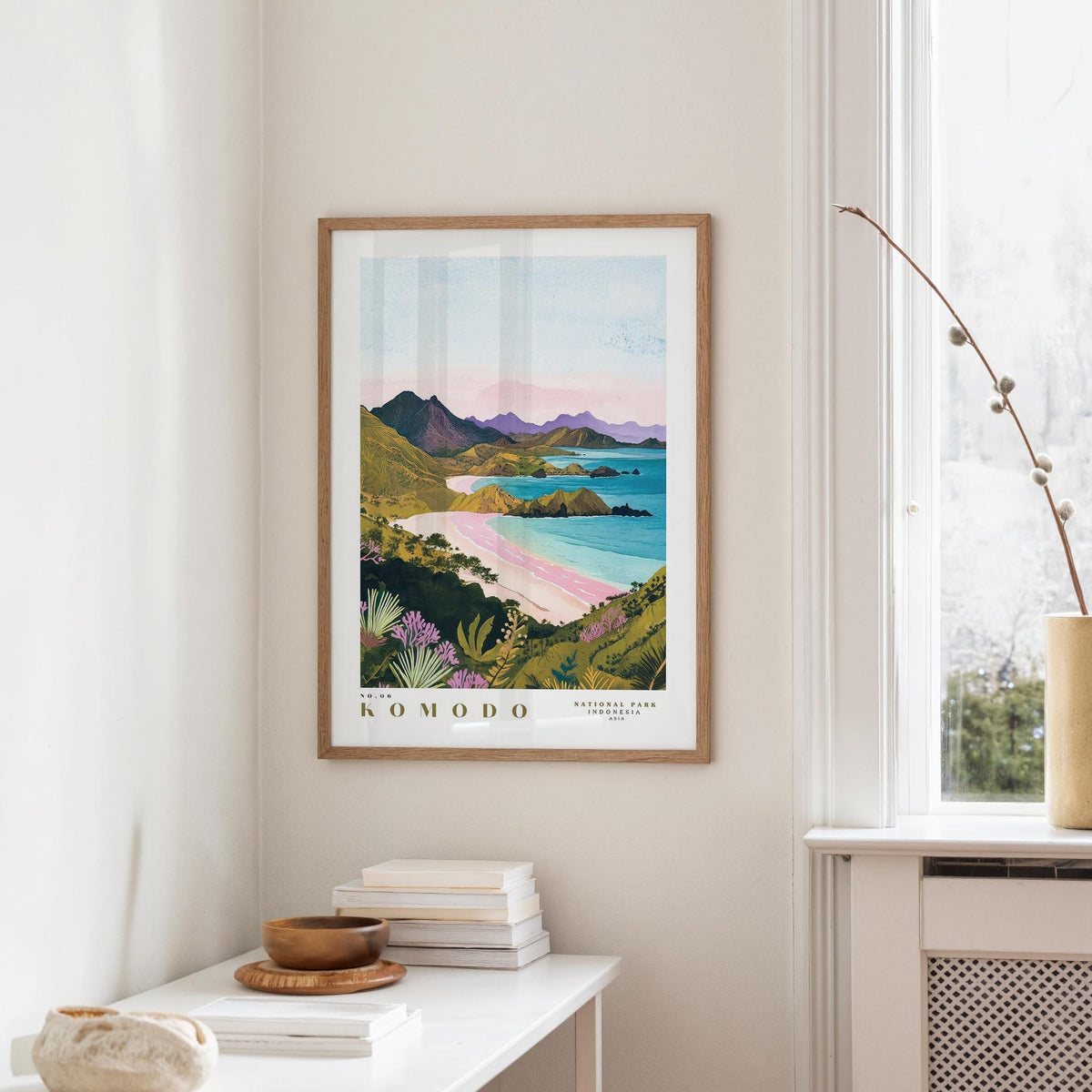 a framed poster of a scenic landscape with mountains, a beach, and a body of water, hanging on a wall in a room with a window and a small table with books and a vase.