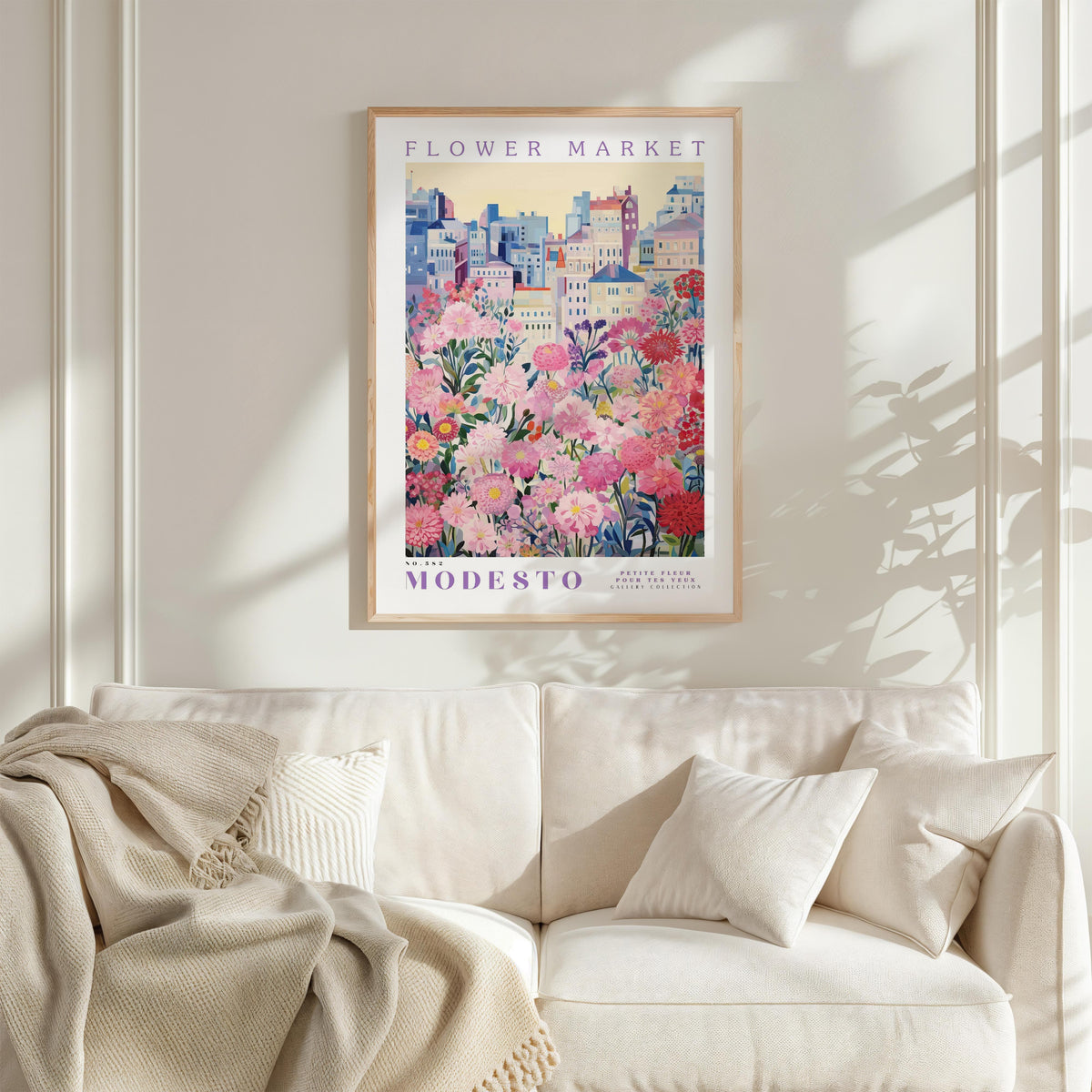 a framed poster of a cityscape with a flower garden in the foreground, hanging on a wall above a white couch with pillows.