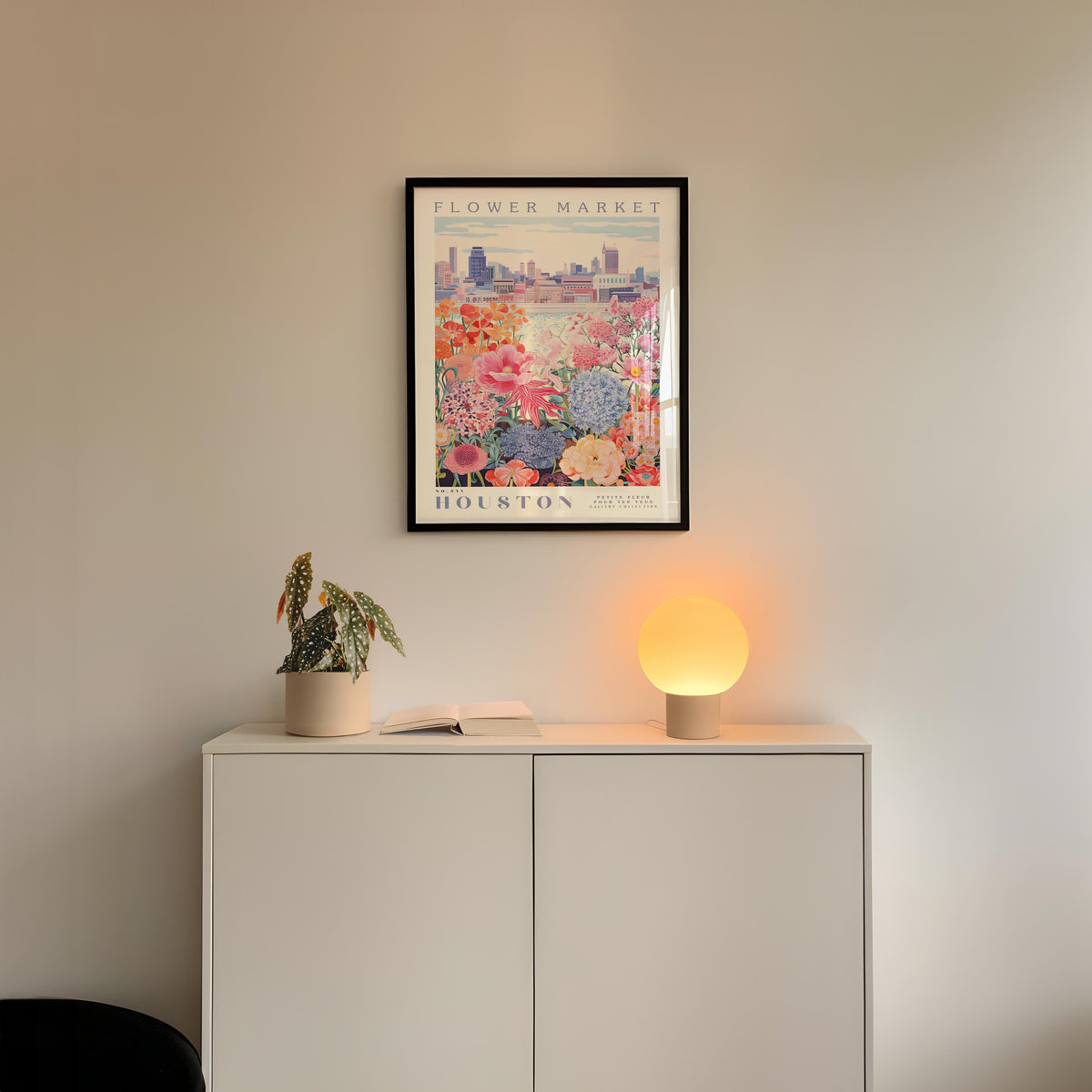 a white cabinet with a potted plant and a lamp on top, set against a wall with a framed poster of a cityscape above it.