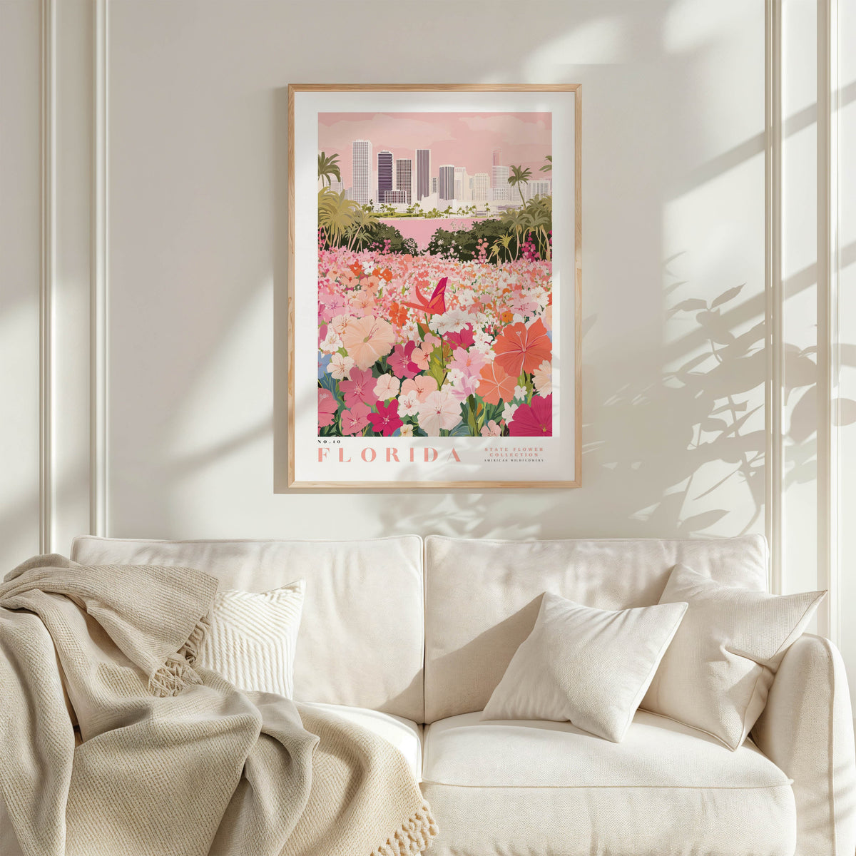 a framed poster of a flower garden with a cityscape in the background, hanging on a wall above a white couch with a blanket draped over it.