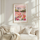 a framed poster of a flower garden with a cityscape in the background, hanging on a wall above a white couch with a blanket draped over it.