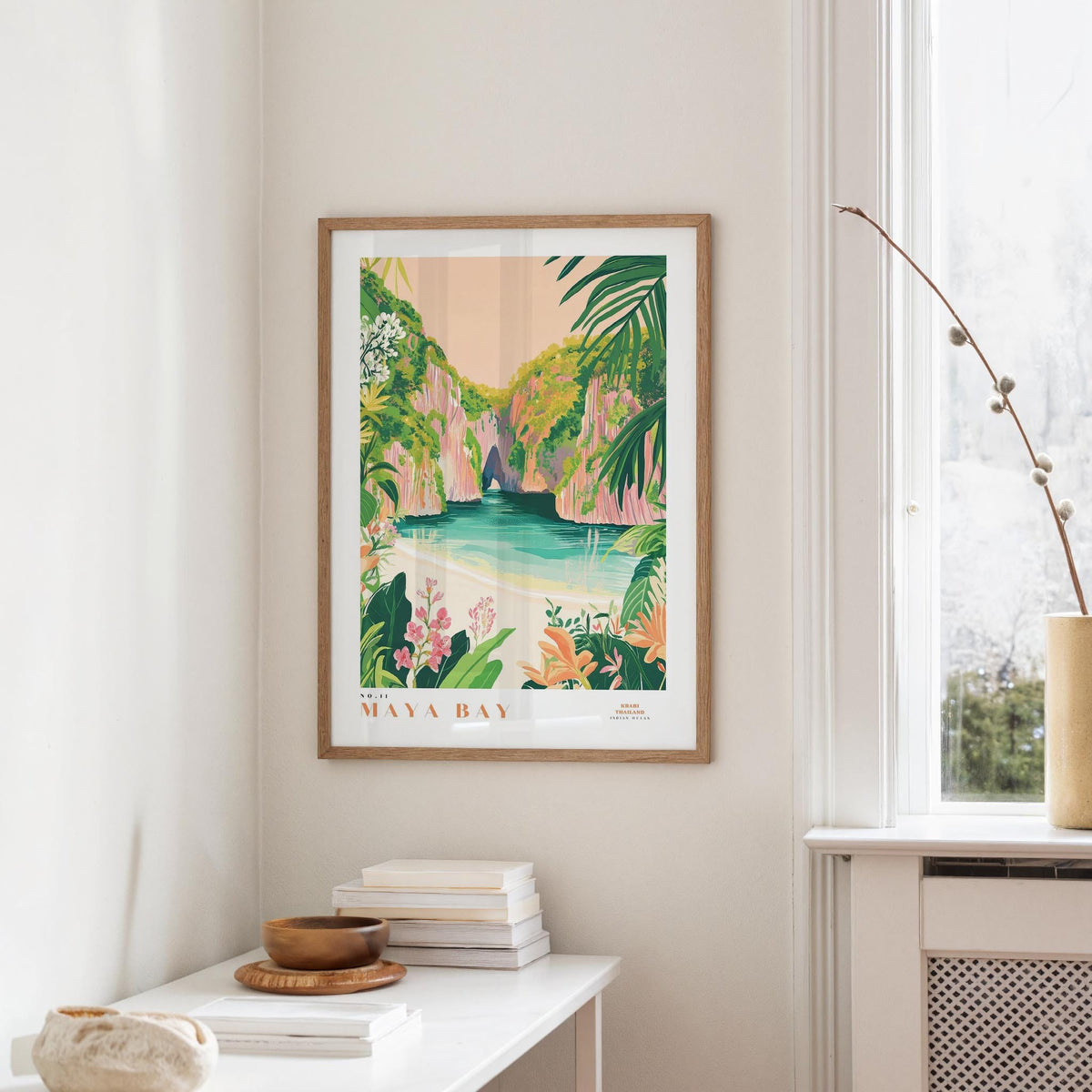 a framed poster of a tropical beach scene, with lush greenery and a turquoise ocean in the background. The poster is hanging on a wall, and there is a small table with a stack of books and a vase on it.