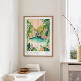 a framed poster of a tropical beach scene, with lush greenery and a turquoise ocean in the background. The poster is hanging on a wall, and there is a small table with a stack of books and a vase on it.