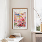 a framed poster of a colorful floral scene hanging on a wall, with a window and a small table with books and a vase nearby.