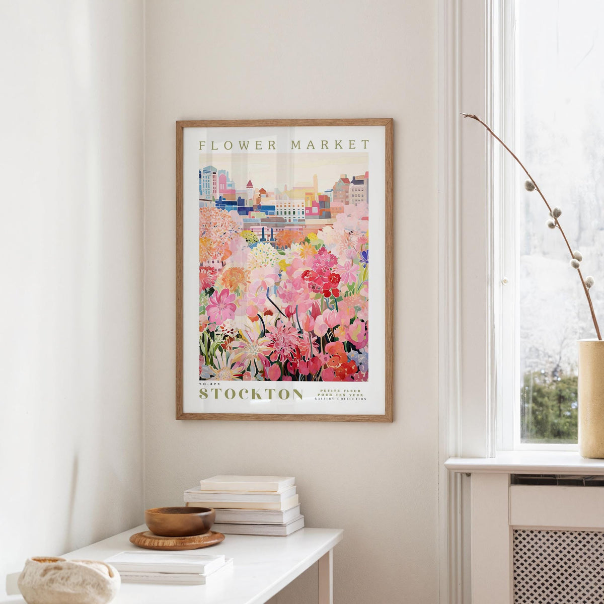 a framed poster of a colorful floral scene hanging on a wall, with a window and a small table with books and a vase nearby.