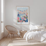 a bedroom with a bed, a woven chair, and a framed poster of a colorful floral scene hanging on the wall.