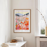 a framed poster of a colorful floral scene hanging on a wall, with a white table and a vase of flowers in the foreground.