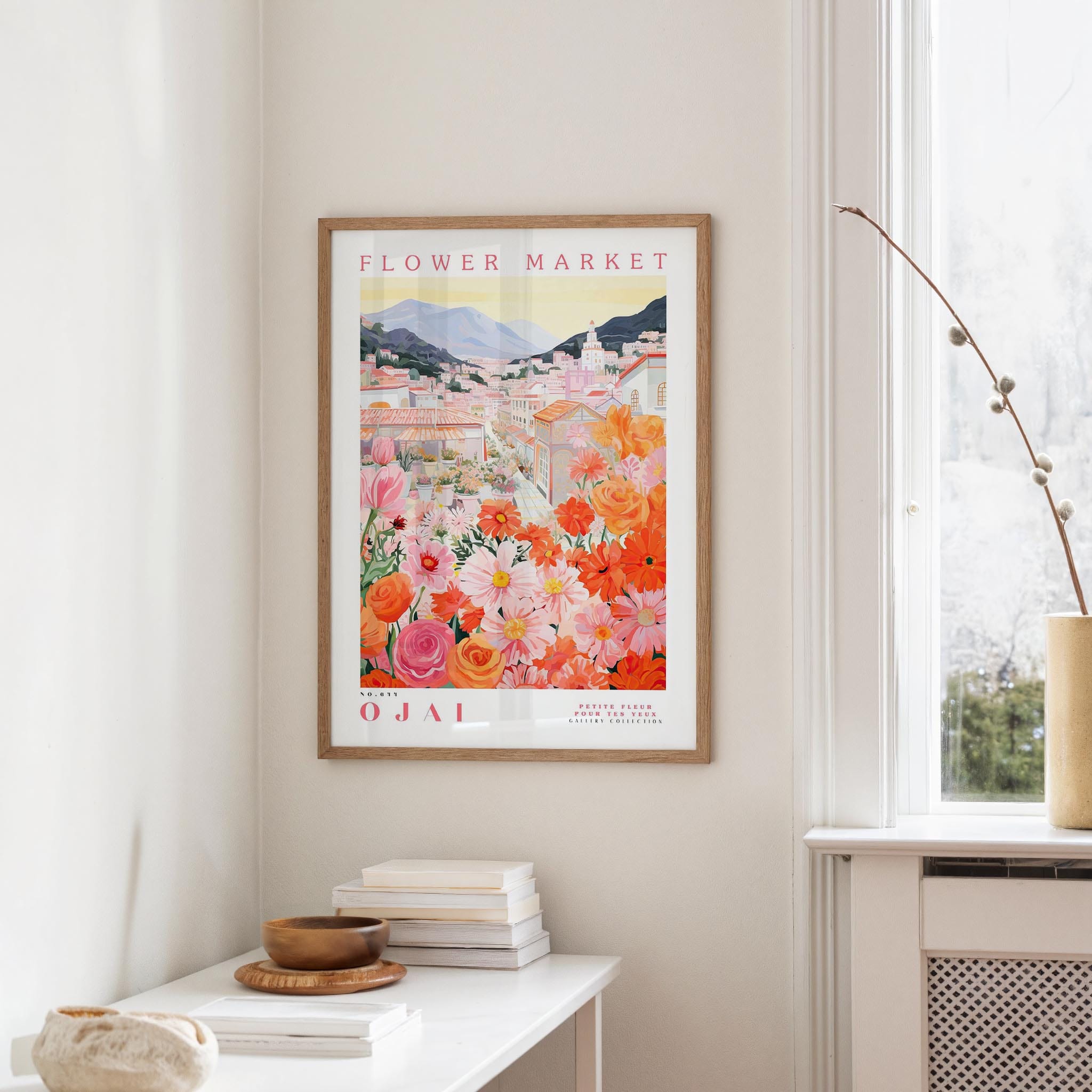 a framed poster of a colorful floral scene hanging on a wall, with a white table and a vase of flowers in the foreground.