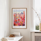 a framed poster of a colorful floral scene hanging on a wall, with a white table and a vase of flowers in the foreground.