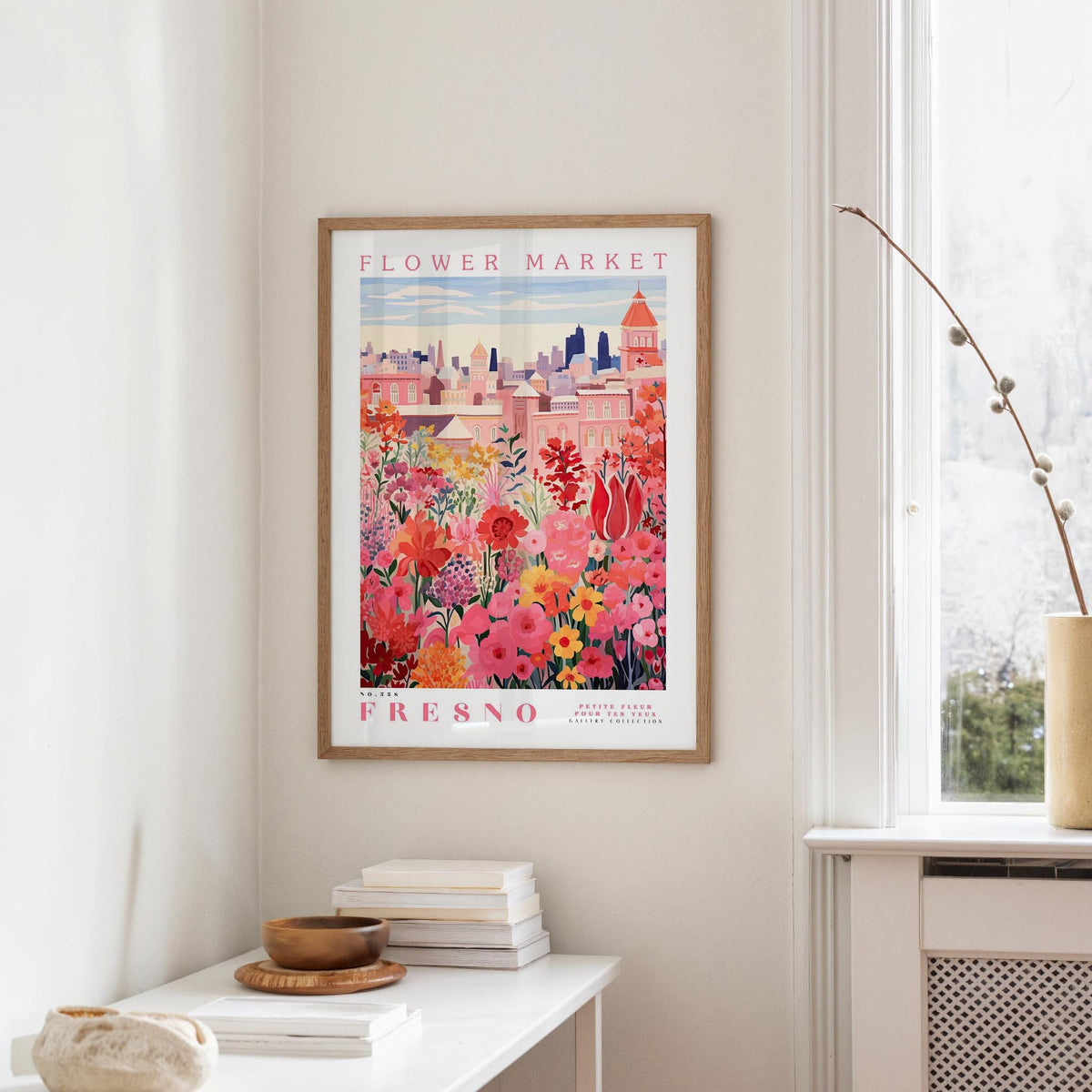 a framed poster of a colorful floral scene hanging on a wall, with a white table and a vase of flowers in the foreground.