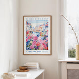 a framed poster of a colorful floral scene hanging on a wall, with a white table and a vase of flowers in the foreground.