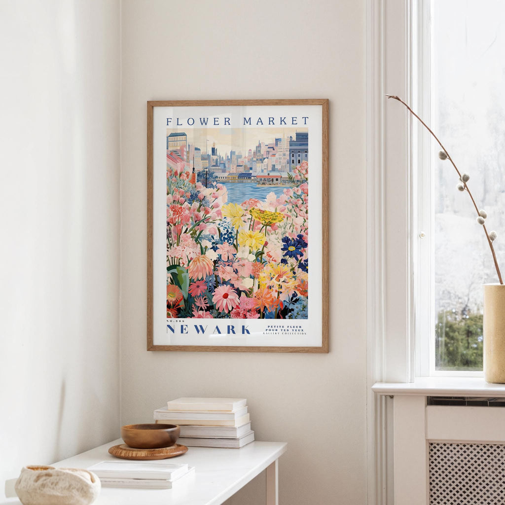 a framed poster of a colorful floral scene hanging on a wall, with a window and a small table with books and a vase in the foreground.