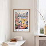 a framed poster of a colorful floral scene hanging on a wall, with a window and a small table with books and a vase in the foreground.