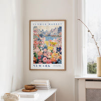 a framed poster of a colorful floral scene hanging on a wall, with a window and a small table with books and a vase in the foreground.
