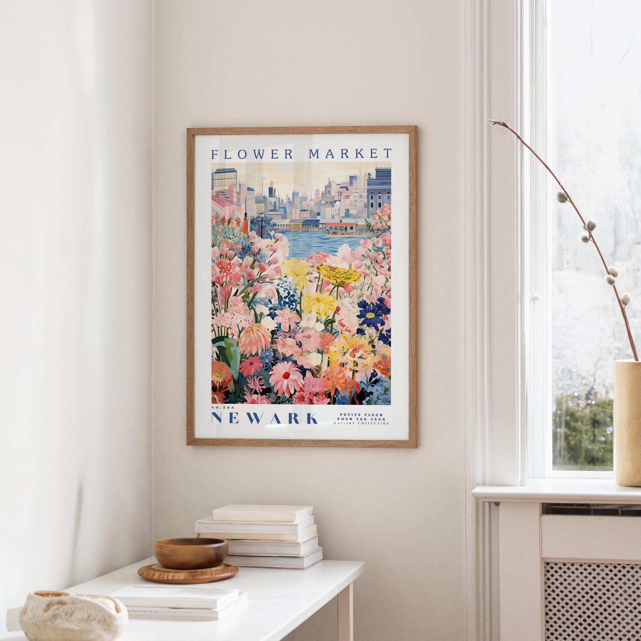 a framed poster of a colorful floral scene hanging on a wall, with a window and a small table with books and a vase in the foreground.