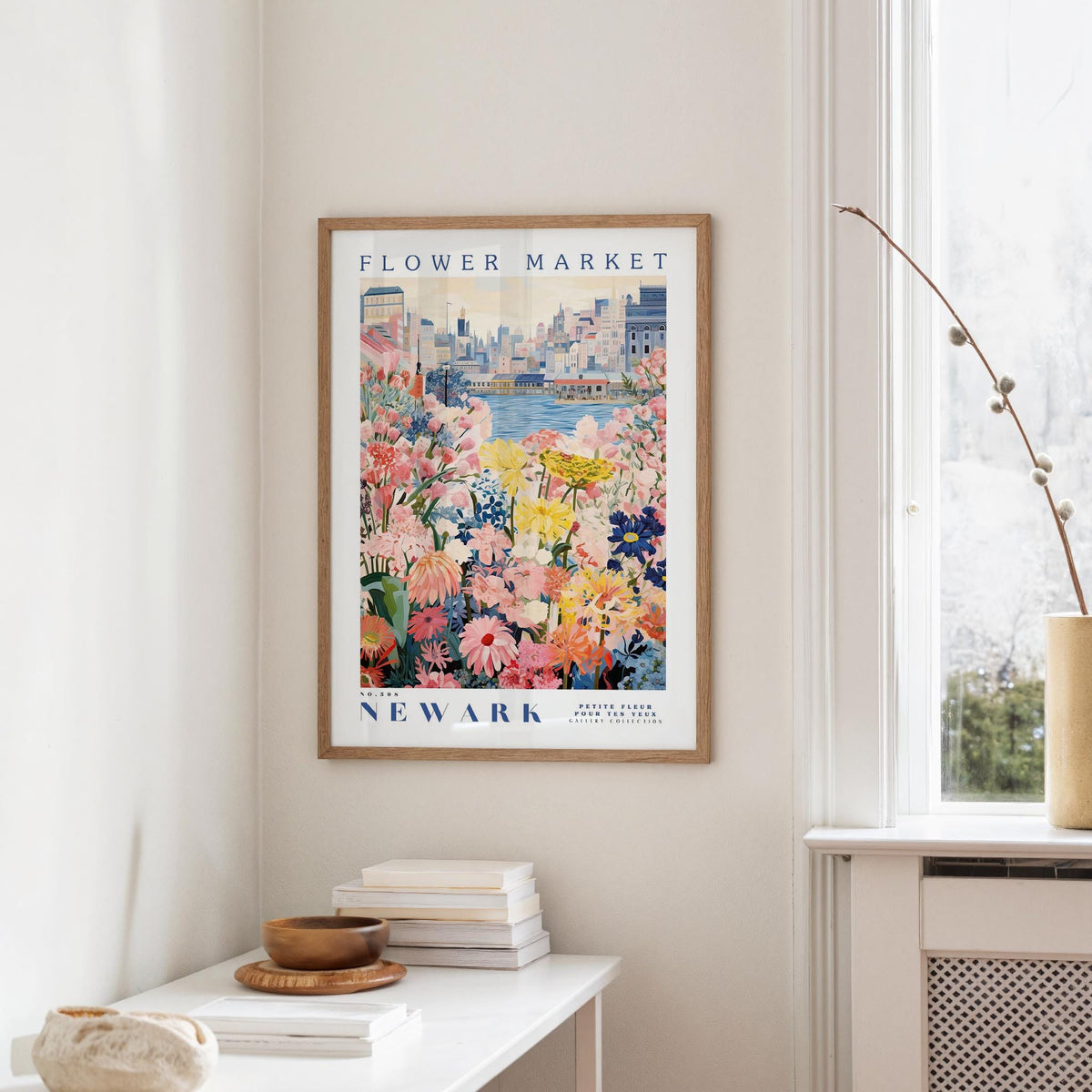a framed poster of a colorful floral scene hanging on a wall, with a window and a small table with books and a vase in the foreground.