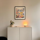 a white cabinet with a potted plant and a lamp on top, set against a wall with a framed poster of a cityscape above it.
