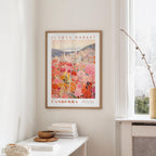 a framed poster of a cityscape with a flower market in the background, hanging on a wall in a room with a window and a small table with books and a vase.