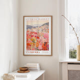 a framed poster of a cityscape with a flower market in the background, hanging on a wall in a room with a window and a small table with books and a vase.