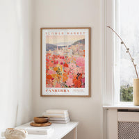a framed poster of a cityscape with a flower market in the background, hanging on a wall in a room with a window and a small table with books and a vase.