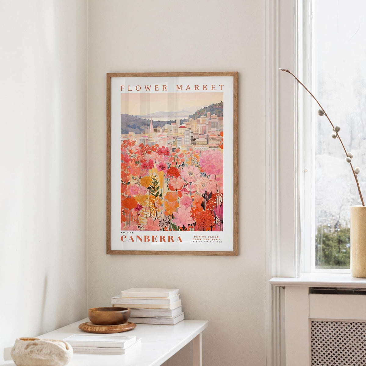 a framed poster of a cityscape with a flower market in the background, hanging on a wall in a room with a window and a small table with books and a vase.