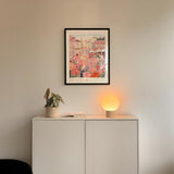 a white cabinet with a potted plant and a lamp on top, set against a white wall. Above the cabinet, there is a framed poster of a cityscape with a pink and orange color scheme.