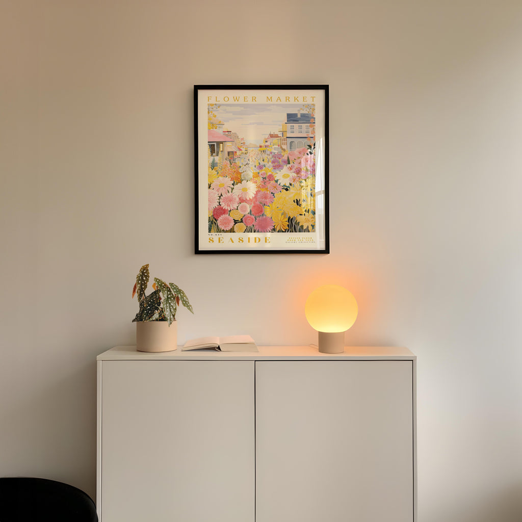 a white cabinet with a potted plant and a lamp on top, set against a wall with a framed poster of a flower market above it.