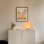 a white cabinet with a potted plant and a lamp on top, set against a wall with a framed poster of a flower market above it.