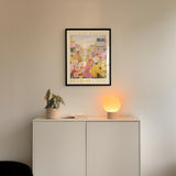 a white cabinet with a potted plant and a lamp on top, set against a wall with a framed poster of a flower market above it.