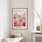 a framed poster of a cityscape with a flower market in the background, hanging on a wall in a room with a window and a small table with books and a vase.