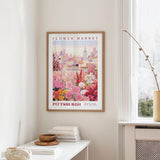 a framed poster of a cityscape with a flower market in the background, hanging on a wall in a room with a window and a small table with books and a vase.