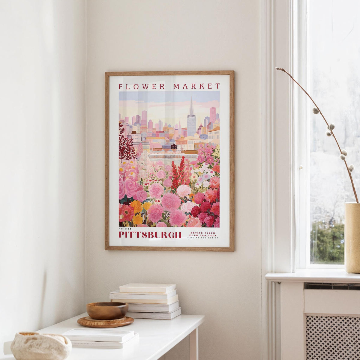 a framed poster of a cityscape with a flower market in the background, hanging on a wall in a room with a window and a small table with books and a vase.
