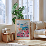 a living room with a large framed poster of a flower market in Corfu, Greece, placed on the floor in front of a window. The poster has a pink and green floral design and the text "FLOWER MARKET" and "CORFU" printed on it. The room also includes a white sofa, a wooden side table with books, and a potted plant.