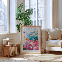 a living room with a large framed poster of a flower market in Corfu, Greece, placed on the floor in front of a window. The poster has a pink and green floral design and the text "FLOWER MARKET" and "CORFU" printed on it. The room also includes a white sofa, a wooden side table with books, and a potted plant.