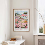 a framed poster of a colorful garden scene hanging on a wall, with a white table and a vase of flowers in the foreground.