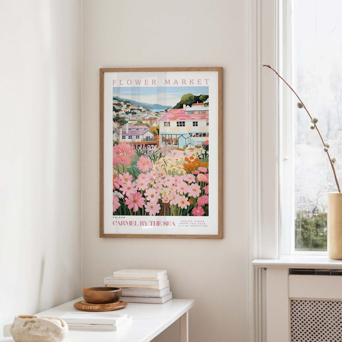 a framed poster of a colorful garden scene hanging on a wall, with a white table and a vase of flowers in the foreground.