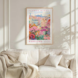 a framed poster of a colorful floral scene hanging on a wall above a white couch with a blanket draped over it.