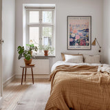 a cozy bedroom with a large bed, a wooden nightstand, and a window with a plant on the sill. Above the bed, there is a framed poster of a flower market.