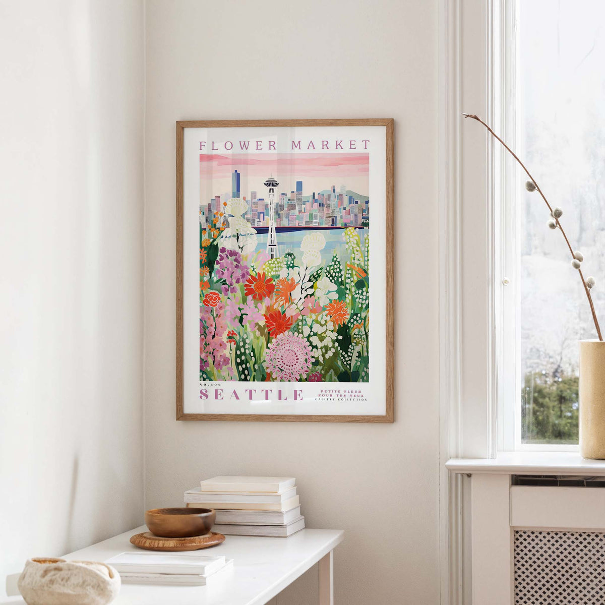 a framed poster of a colorful floral scene hanging on a wall, with a window and a small table with books and a vase in the foreground.
