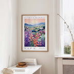 a framed poster of a colorful floral landscape hanging on a wall, with a window and a table with books and a vase in the foreground.