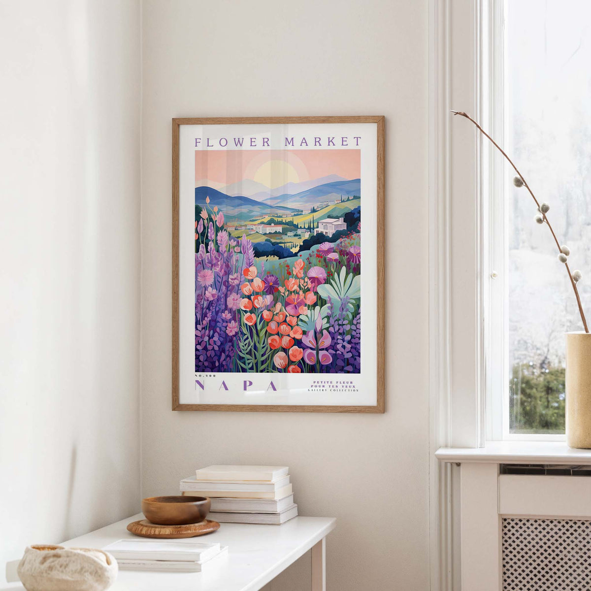 a framed poster of a colorful floral landscape hanging on a wall, with a window and a table with books and a vase in the foreground.