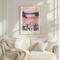 a framed poster of a scenic landscape with a lake and mountains, hanging on a wall above a white couch with a blanket draped over it.