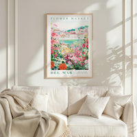 a framed poster of a colorful garden scene hanging on a wall above a white couch with a blanket draped over it.