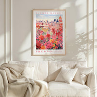 a framed poster of a vibrant floral scene hanging on a wall above a white couch with a blanket draped over it.
