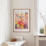 a framed poster of a colorful floral scene hanging on a wall, with a window and a small table with books and a vase in the foreground.