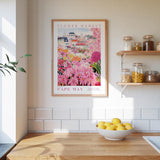 a kitchen scene with a framed poster of a colorful flower market in Cape May, New Jersey, hanging on the wall above a wooden counter. On the counter, there is a bowl of lemons and a plant.