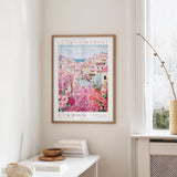 a framed poster of a colorful cityscape with pink flowers hanging on a wall, next to a window and a small table with books and a bowl.