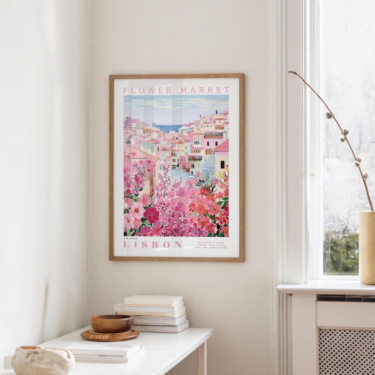 a framed poster of a colorful cityscape with pink flowers hanging on a wall, next to a window and a small table with books and a bowl.