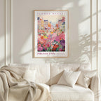 a framed poster of a colorful floral scene hanging on a wall above a white couch with a blanket draped over it.