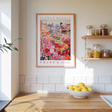 a kitchen scene with a framed poster of a colorful flower market in Frisco, California, hanging on the wall above a wooden counter. On the counter, there is a bowl of lemons and a plant.